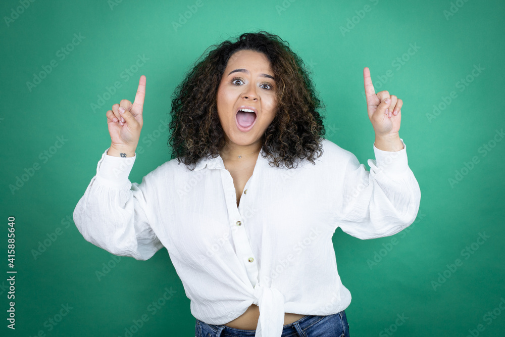 Young african american girl wearing white shirt over green background amazed and surprised looking at the camera and pointing up with fingers and raised arms Young african american girl wearing white shirt over green background amazed and surprised looking at the camera and pointing up with fingers and raised arms