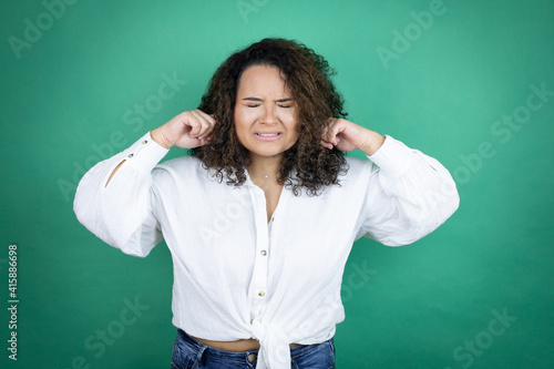 Young african american girl wearing white shirt over green background covering ears with fingers with annoyed expression for the noise of loud music. Deaf concept. © Irene