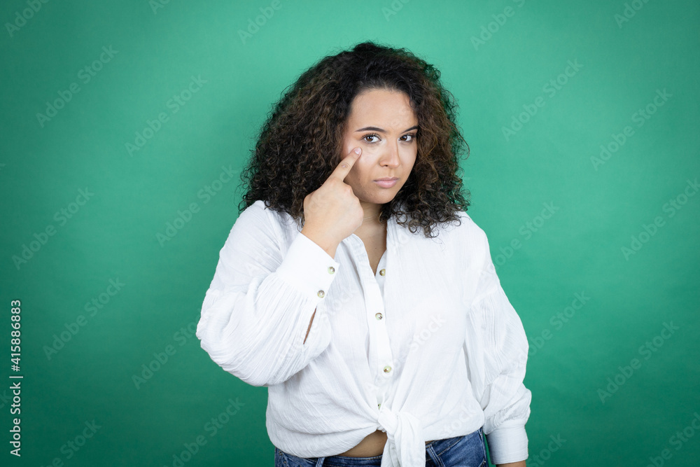 Young african american girl wearing white shirt over green background Pointing to the eye watching you gesture, suspicious expression Young african american girl wearing white shirt over green background Pointing to the eye watching you gesture, suspicious expression