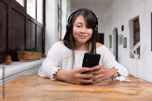 Beautiful happy latina woman with long black hair and wireless headphones listening to music leaning on a wooden table using her cell phone with her hands