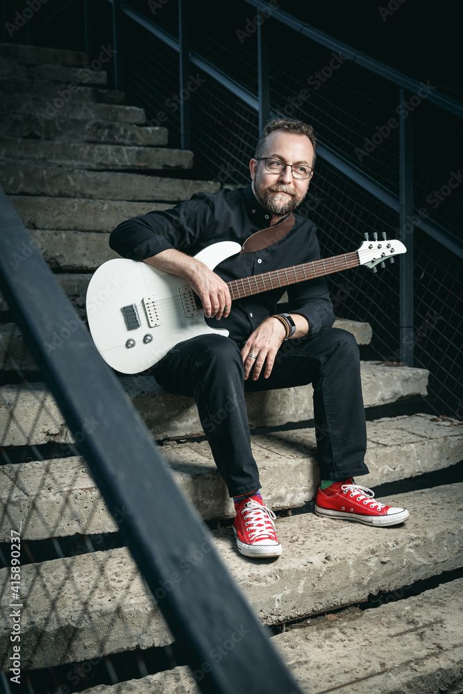 Obraz premium Portrait of a male guitar player with electric guitar wearing black clothing and red sneackers sitting on a stairs
