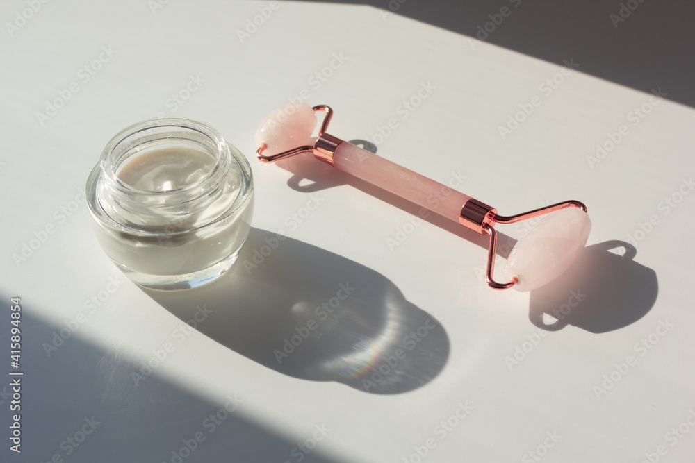 Concept of minimalist skincare routine (skinimalism). Closeup of moisturizer and rose quartz roller with contrasting shadows.