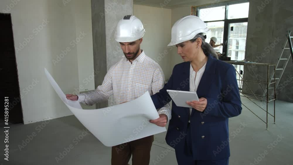 Architects in protective helmets holding blueprint and tablet pc while walking around apartment under renovation. Professional designers working on apartment layout among empty space and bare walls Architects in protective helmets holding blueprint and tablet pc while walking around apartment under renovation. Professional designers working on apartment layout among empty space and bare walls