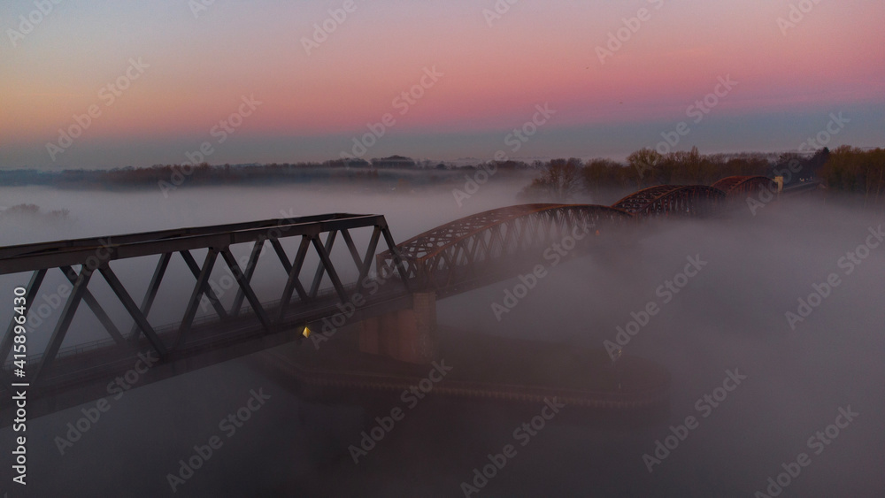 Obraz premium Bridge at Sunrise with a red sky in the Horizon . The picture was taken by a drone. the River unter the Bridge called Rhein. scary fog is around the bridge.