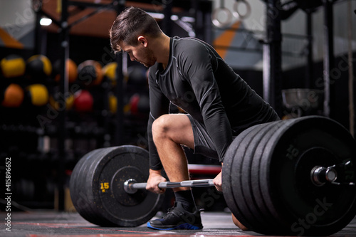 Athlete standing on his knee preparing to make deadlift at gym, young caucasian man in black sportswear engaged in bodybuilding, concentrated on weightlifting. sport, cross fit