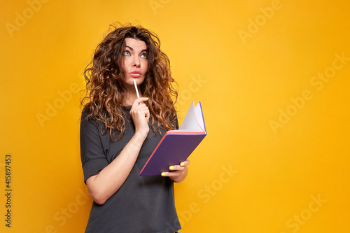 The curly-haired woman with the purple diary and the white pencil looked thoughtful and looked away. She's wearing a dark gray T-shirt. It stands on a yellow solid background.