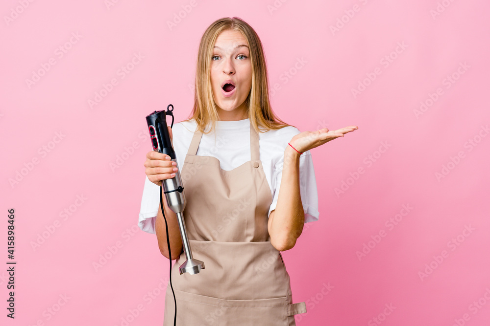 Young russian cook woman holding an electric mixer isolated surprised and shocked. Young russian cook woman holding an electric mixer isolated surprised and shocked.