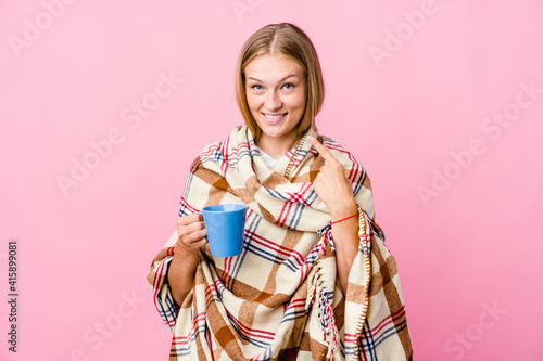 Young russian woman wrapped in a blanket drinking coffee smiles  pointing fingers at mouth.