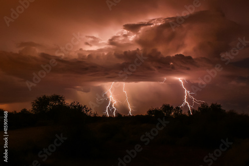Lightning strike during summer thunderstorm, in Arizona's Sonoran Desert. Three bolts extend down from the clouds, which are backlit by the discharge. Silhouettes of brush in the foreground. 