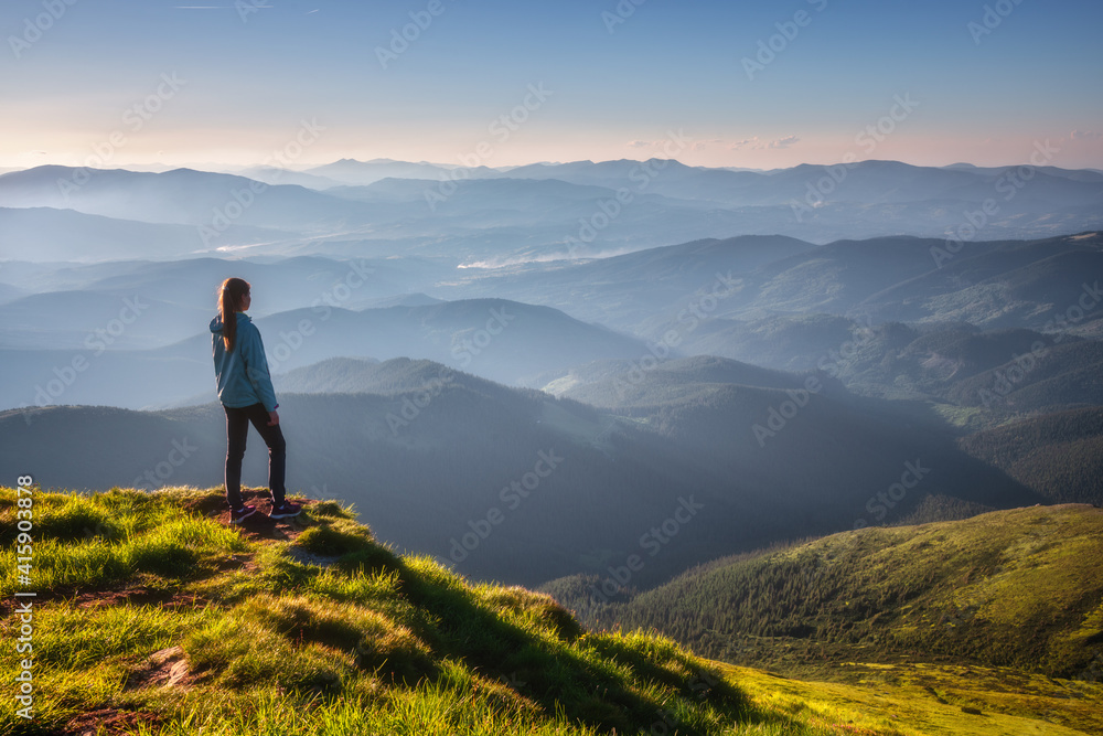 Girl on mountain peak with green grass looking at beautiful mountain valley in fog at sunset in summer. Landscape with sporty young woman, foggy hills, forest, sky. Travel and tourism. Hiking - obrazy, fototapety, plakaty 