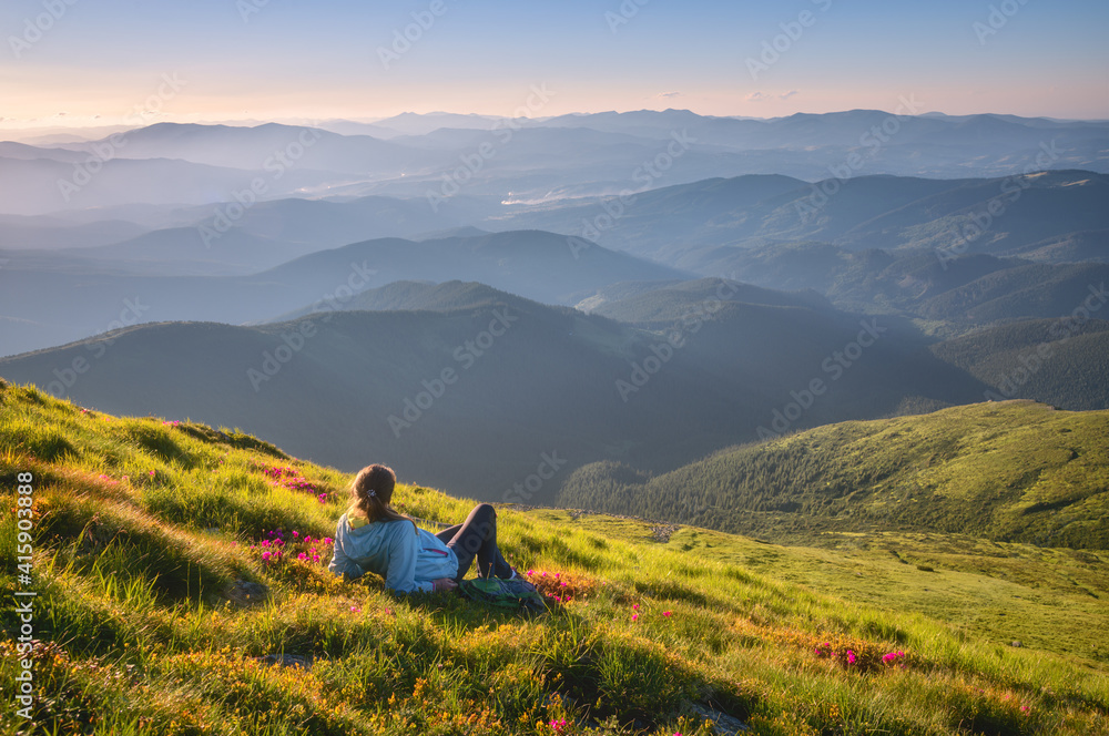 Young woman on the mountain peak with green grass and pink flowers looking at beautiful mountains in fog at sunset in summer. Colorful landscape with lying girl, forest, hills, sky. Travel and tourism