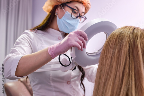 young dermatologist looking at a dermatoscope while examining female head and hair. in a bright cosmetology room, cabinet. photo