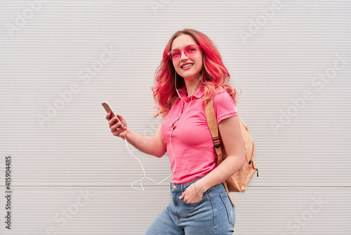Young blogger woman with pink dyed hairs staying near the grey wall and using smartphone