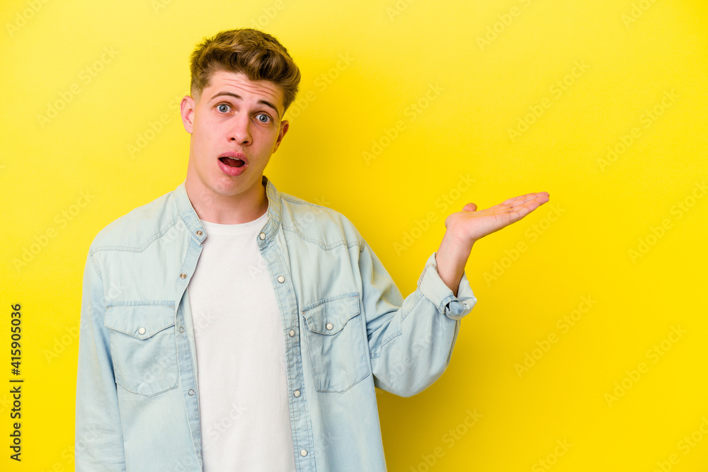 Young caucasian man isolated on yellow background impressed holding copy space on palm. Young caucasian man isolated on yellow background impressed holding copy space on palm.