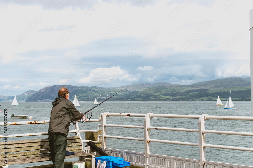Old fisherman with a fishing rod wearing a raincoat and fishing in a pier with sailboats, mountains and cloudy sky in the background