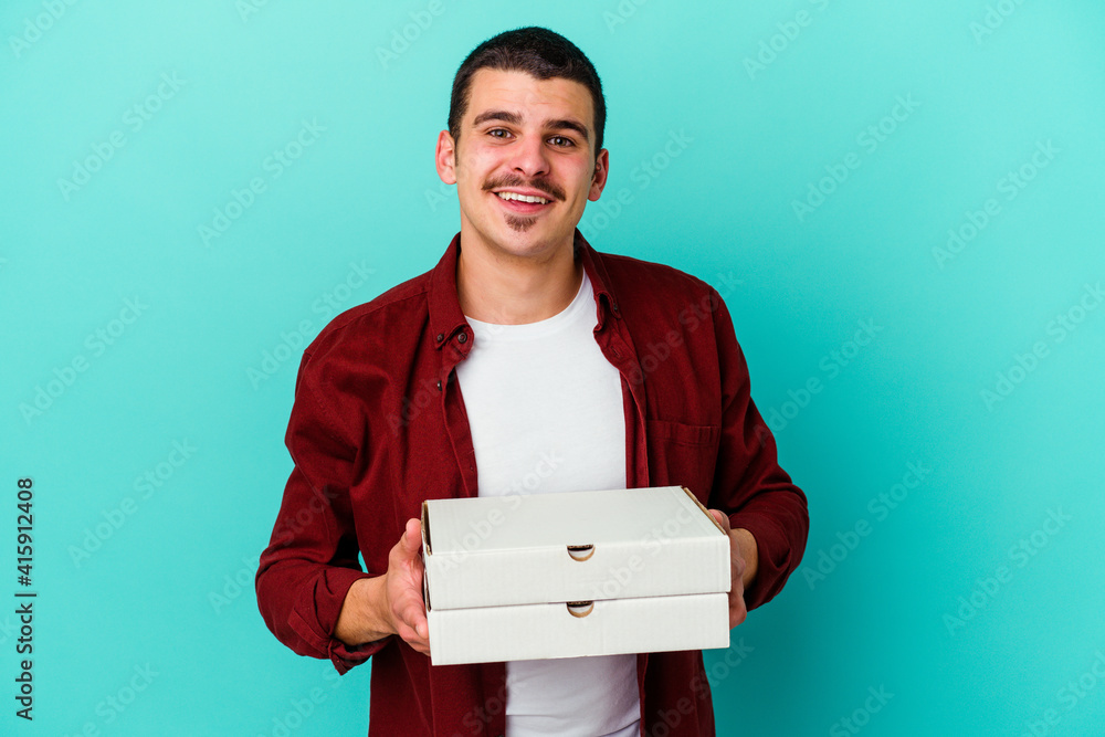 Young caucasian man holding pizzas isolated on blue background happy, smiling and cheerful. Young caucasian man holding pizzas isolated on blue background happy, smiling and cheerful.