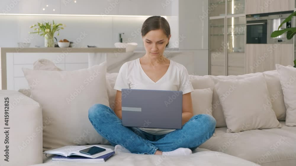 Young woman in casual clothes working on laptop while sitting on sofa at home Young woman in casual clothes working on laptop while sitting on sofa at home
