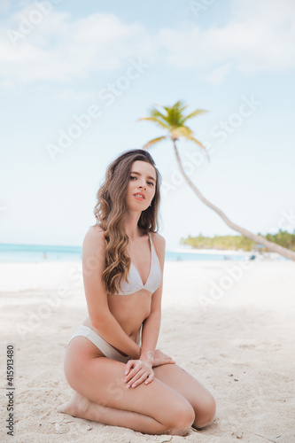 Portrait of young woman in white bikini on tropical beach looking at camera. Beautiful latin girl in swimwear with copy space. Summer vacation and tanning concept.