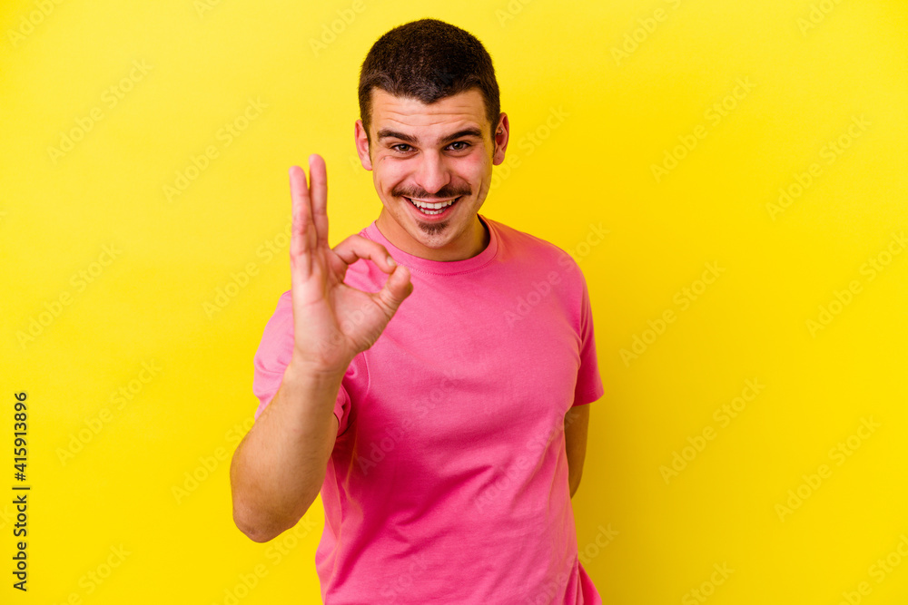 Young caucasian cool man isolated on yellow background winks an eye and holds an okay gesture with hand. Young caucasian cool man isolated on yellow background winks an eye and holds an okay gesture with hand.