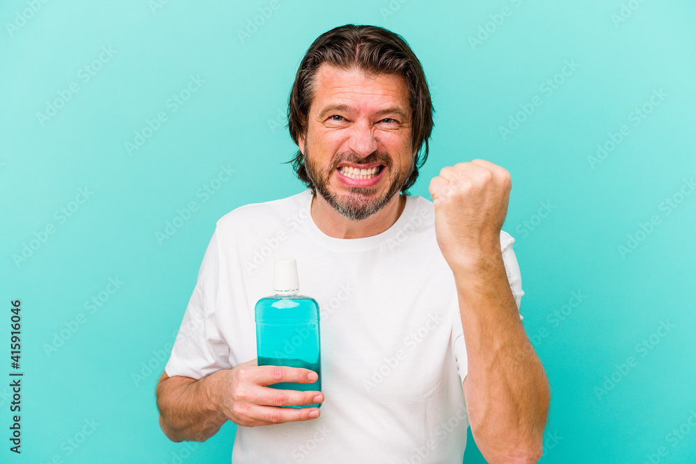 Middle age dutch man sitting holding a mouthwash isolated on blue background showing fist to camera, aggressive facial expression. Middle age dutch man sitting holding a mouthwash isolated on blue background showing fist to camera, aggressive facial expression.