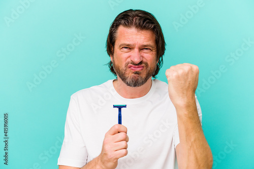Middle age dutch man holding a razor blade isolated on blue background showing fist to camera, aggressive facial expression.
