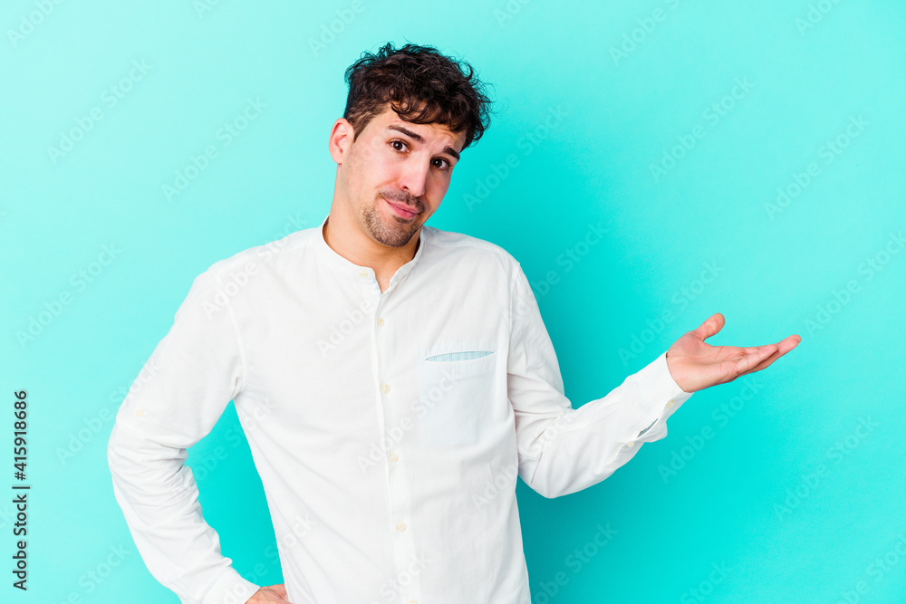 Young caucasian man isolated on blue background doubting and shrugging shoulders in questioning gesture.