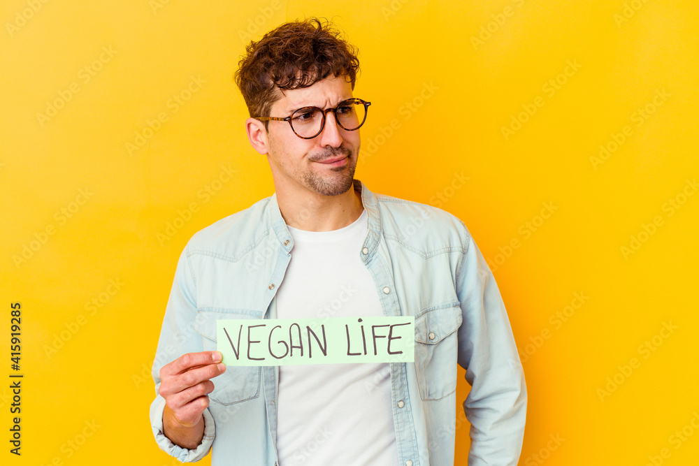 Young caucasian man holding a vegan life placard isolated confused, feels doubtful and unsure. Young caucasian man holding a vegan life placard isolated confused, feels doubtful and unsure.