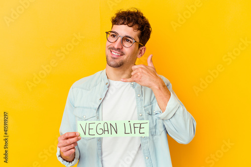 Young caucasian man holding a vegan life placard isolated showing a mobile phone call gesture with fingers.