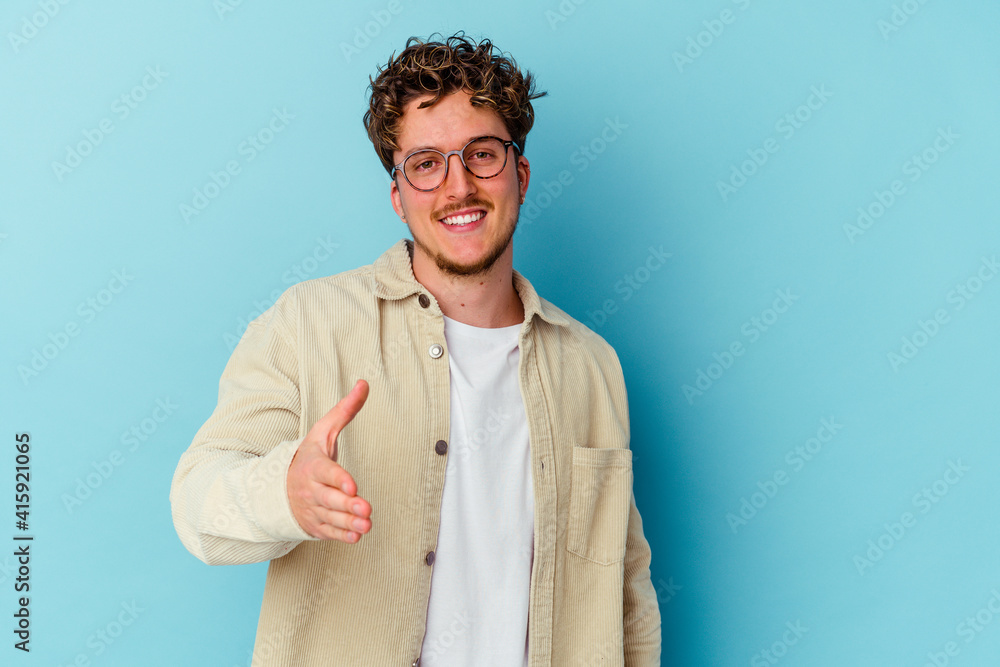 Young caucasian man wearing eyeglasses isolated on blue background stretching hand at camera in greeting gesture. Young caucasian man wearing eyeglasses isolated on blue background stretching hand at camera in greeting gesture.