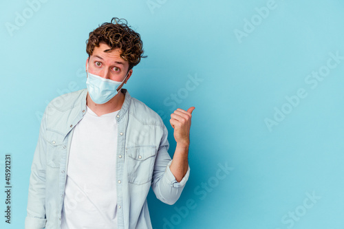 Young caucasian man wearing an antiviral mask isolated on blue background points with thumb finger away, laughing and carefree. © Asier
