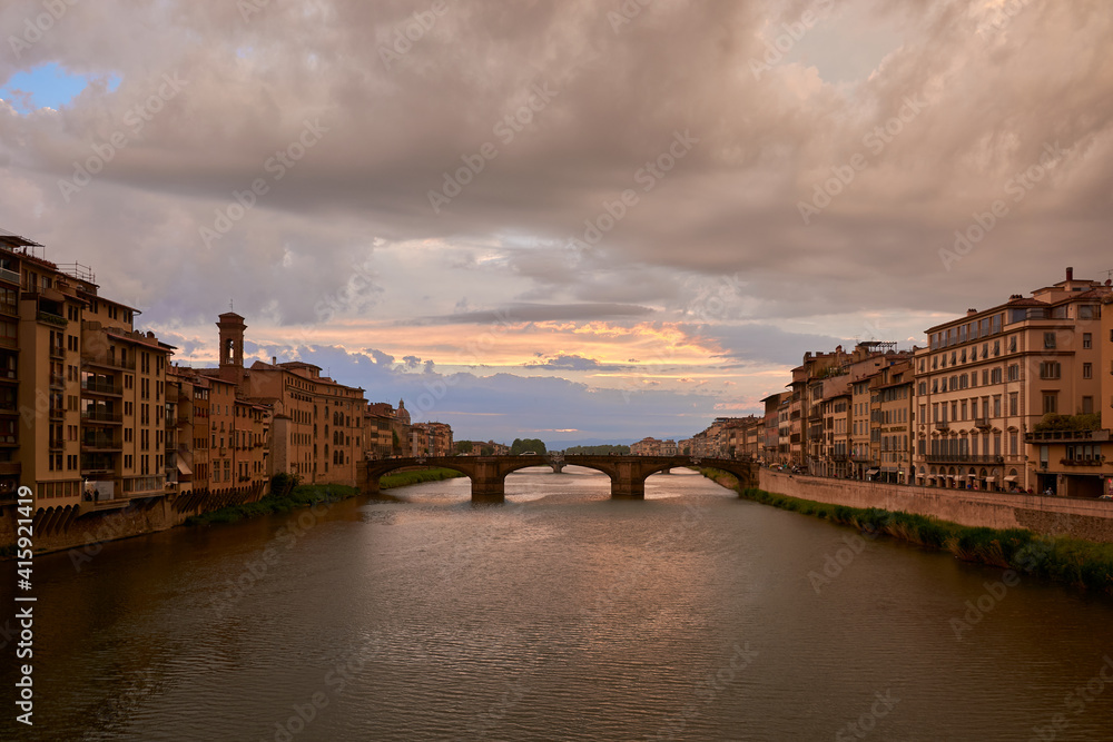 A view of Saint Trinity bridge and the Arno river from Ponte Vecchio, surrounded by picturesque buildings under a beautiful sunset sky on a spring day in Florence, Italy.