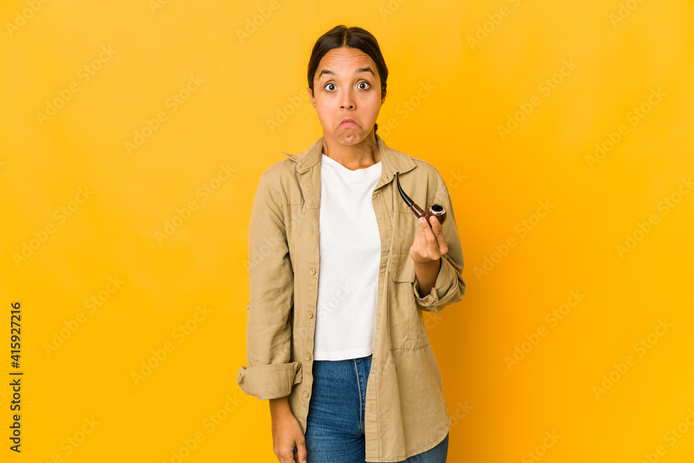 Young hispanic woman holding a smoking pipe shrugs shoulders and open eyes confused. Young hispanic woman holding a smoking pipe shrugs shoulders and open eyes confused.