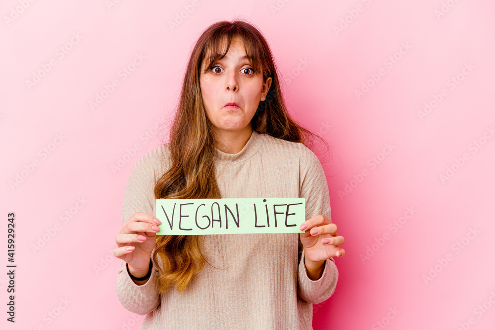 Young caucasian woman holding a vegan life placard isolated shrugs shoulders and open eyes confused. Young caucasian woman holding a vegan life placard isolated shrugs shoulders and open eyes confused.