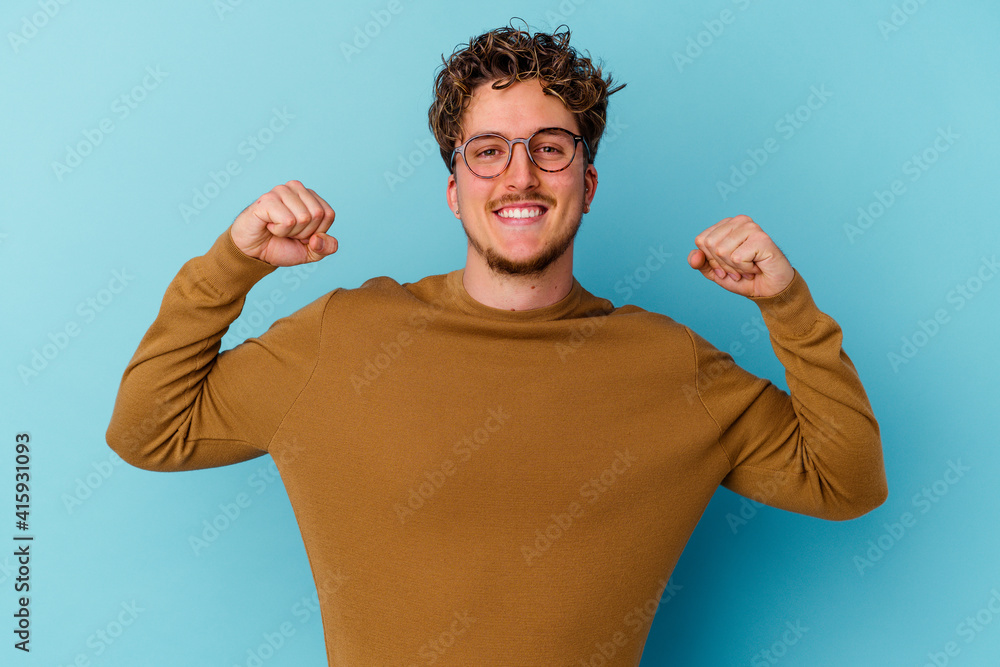 Young caucasian man wearing eyeglasses isolated on blue background showing strength gesture with arms, symbol of feminine power Young caucasian man wearing eyeglasses isolated on blue background showing strength gesture with arms, symbol of feminine power