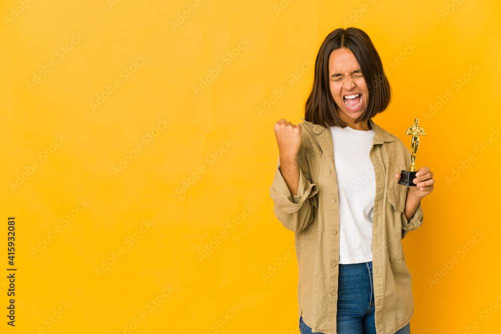 Fototapeta premium Young hispanic woman holding an award statuette raising fist after a victory, winner concept.