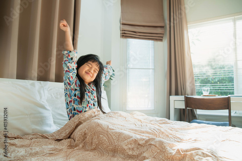 Asian child girl enjoys sunny morning. Good time at home. Kid wakes up from sleep. child girl wakes up in morning in bed and stretches by window. morning awakening little child girl in bed.