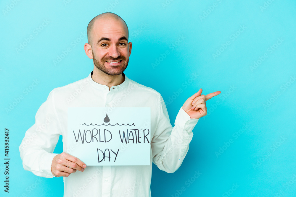 Young caucasian bald man celebrating world water day isolated on blue background smiling and pointing aside, showing something at blank space. Young caucasian bald man celebrating world water day isolated on blue background smiling and pointing aside, showing something at blank space.
