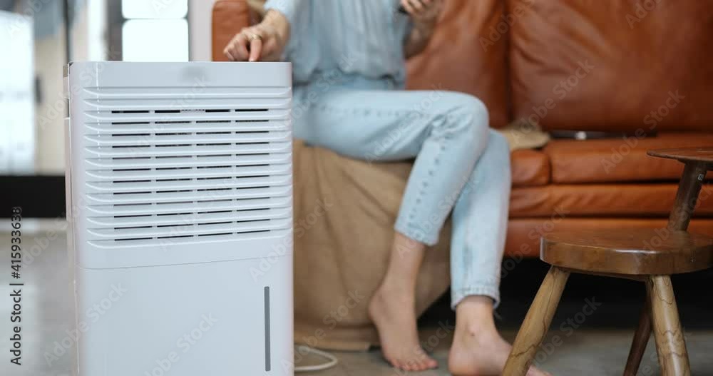 Woman switching on portable air conditioner while sitting on a couch at home. Enjoying fresh and clean air at home. Home appliances for cleaning and air conditioning