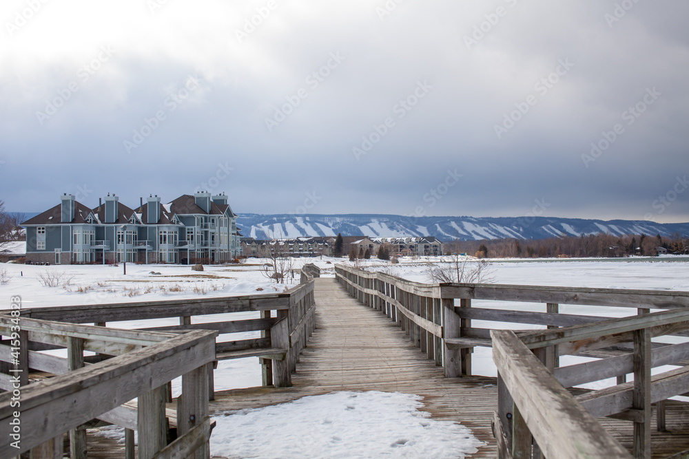A wooden bridge in Lighthouse Point, Collingwood, crosses a portion of Georgian Bay while overlooking the Blue Mountain Ski hills from a distance, with the frozen water beside it