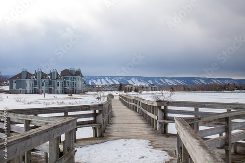 A wooden bridge in Lighthouse Point  Collingwood  crosses a portion of Georgian Bay while overlooking the Blue Mountain Ski hills from a distance  with the frozen water beside it