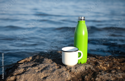 Modern green thermos bottle and cup on beach. Space for text