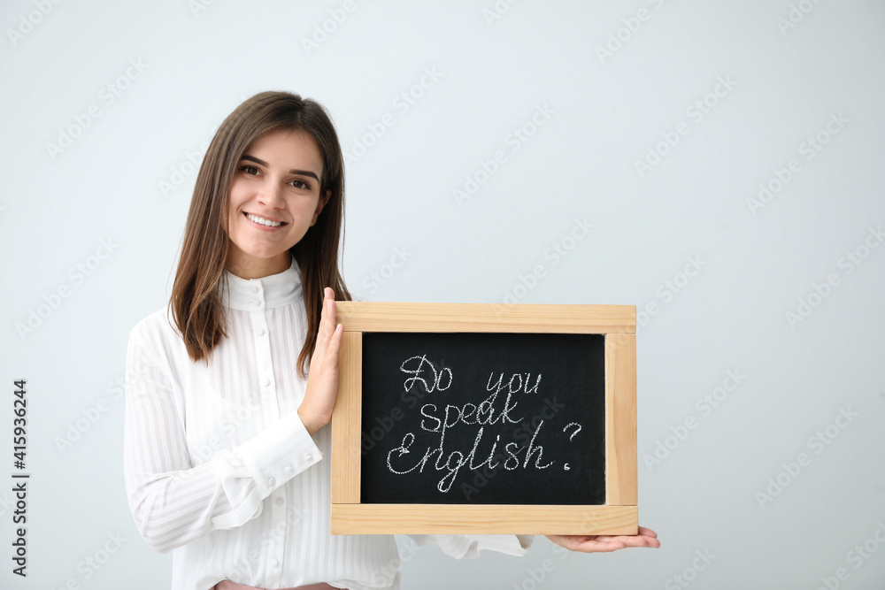Young female teacher holding chalkboard with words DO YOU SPEAK ENGLISH? on light background Young female teacher holding chalkboard with words DO YOU SPEAK ENGLISH? on light background