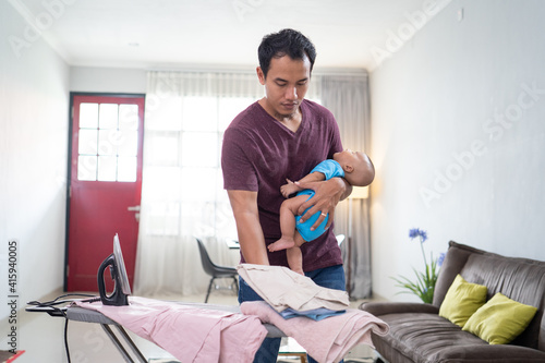 portrait of asian father ironing his clothes while holding his infant baby on his hand