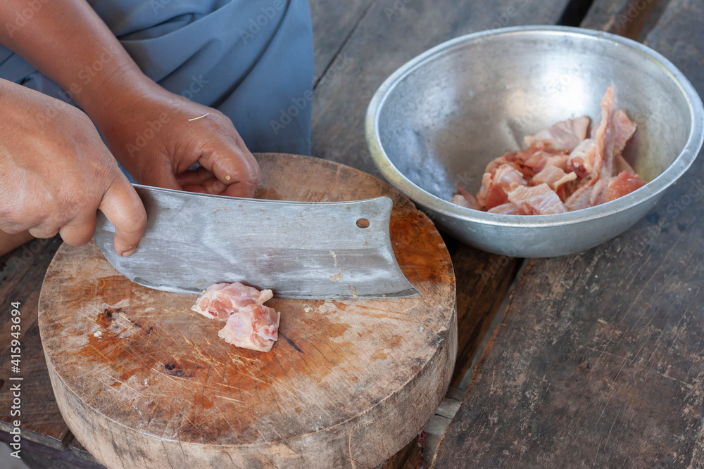 The cook's hand holds the knife chopping chicken on a wooden cutting board to cook in a country kitchen.