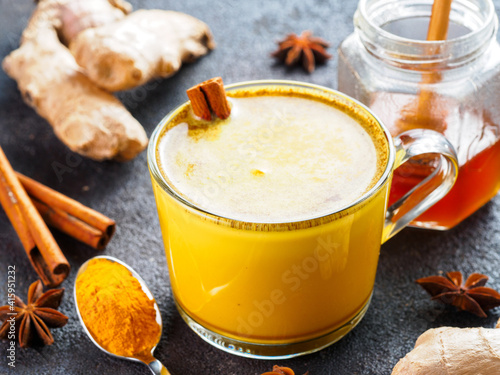 Healthy drink golden turmeric latte in glass cup.Gold milk with turmeric,ginger root,cinnamon sticks,dried turmeric powder and honey over black cement background.Detox turmeric tea and ingredients.