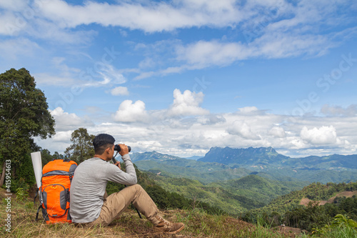 Young man with backpack and holding a binoculars looking on top of mountain at sunset