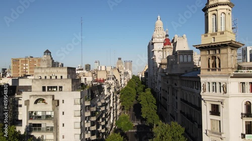 Aerial dolly in flying over Avenida de Mayo surrounded by trees with Barolo Palace tower and Buenos Aires buildings at sunset photo
