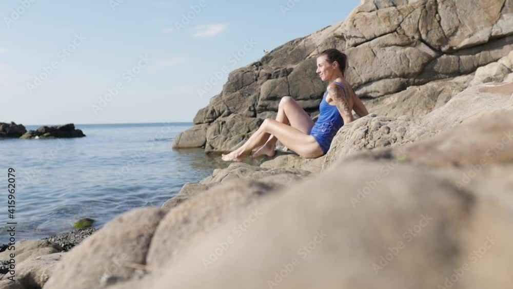 young woman in a swimsuit sitting on a stone beach with the sea. beautiful skinny woman with blond hair enjoys life young woman in a swimsuit sitting on a stone beach with the sea. beautiful skinny woman with blond hair enjoys life