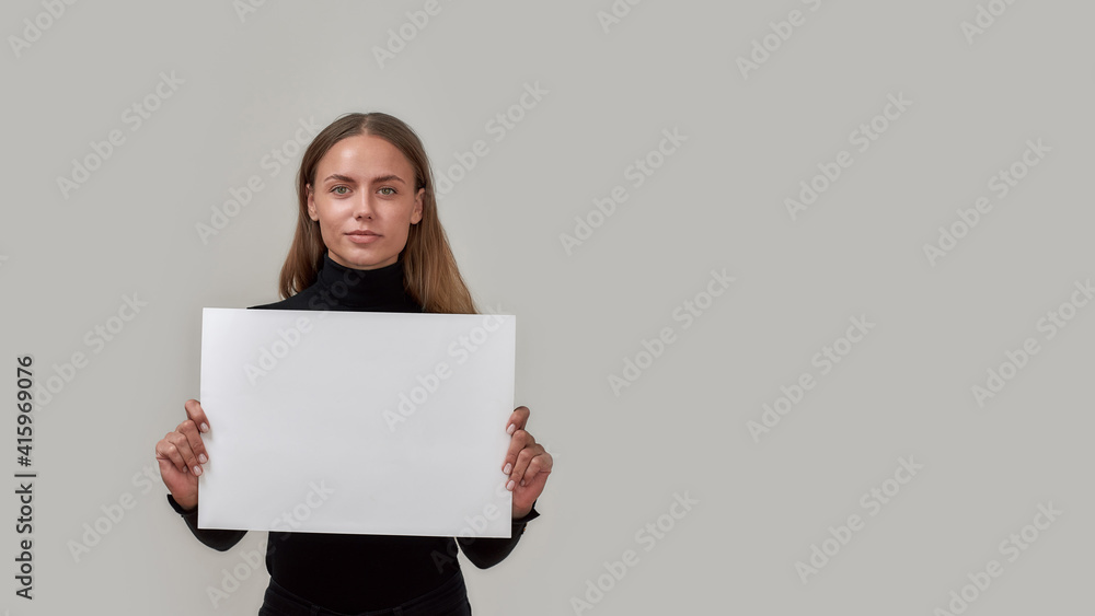 Portrait of young caucasian woman looking at camera, holding white blank banner in front of her while standing isolated over gray background Portrait of young caucasian woman looking at camera, holding white blank banner in front of her while standing isolated over gray background