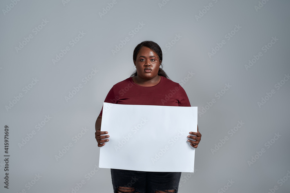 Serious plus size young african american woman in casual clothes looking at camera, holding blank white banner in front of her, posing isolated over gray background Serious plus size young african american woman in casual clothes looking at camera, holding blank white banner in front of her, posing isolated over gray background